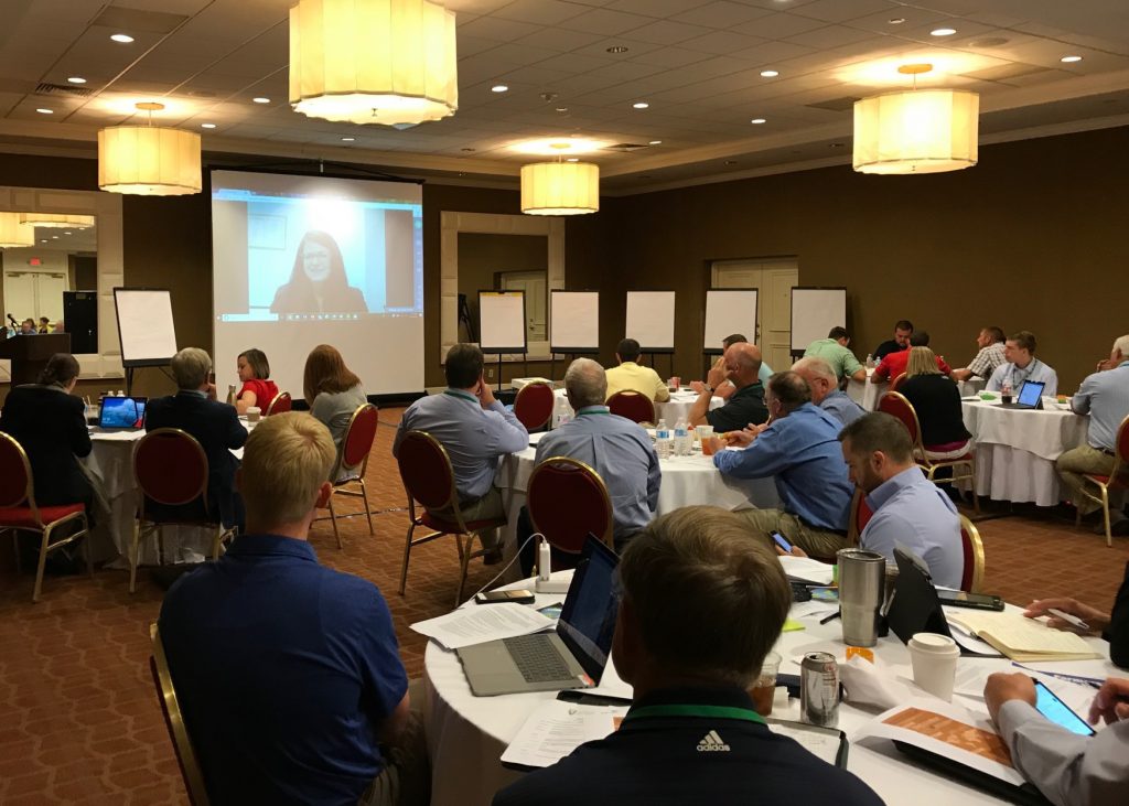 A room of people listening to a speech on a projector