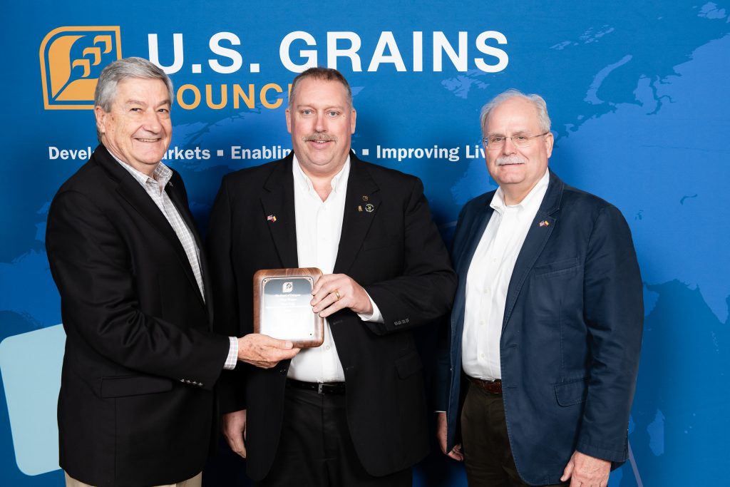 Chad Willis Recognition- 3 men standing in front of Council banner, man in the center holding recognition plaque