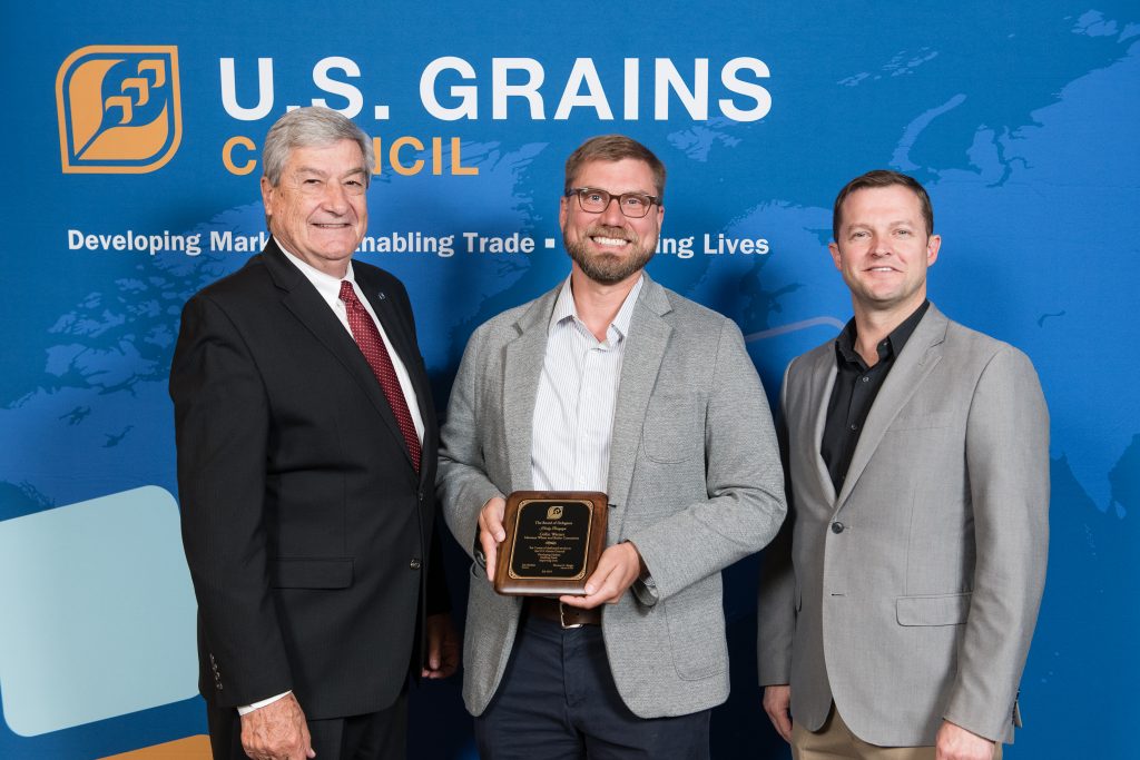 Collin Watters recognition- 3 men standing in front of council banner, man in the center holding recognition plaque