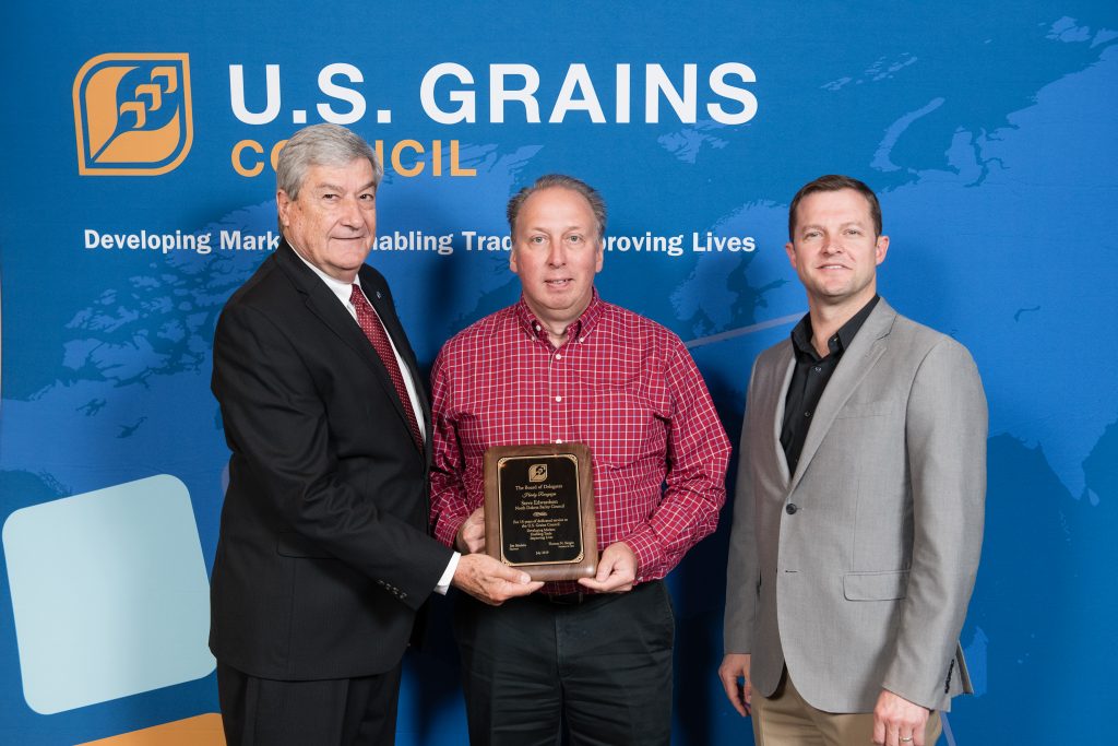 Steve Edwardson Recognition- 3 men standing in front of the Grains & BioProducts Council backdrop, man in the middle holding a recognition plaque